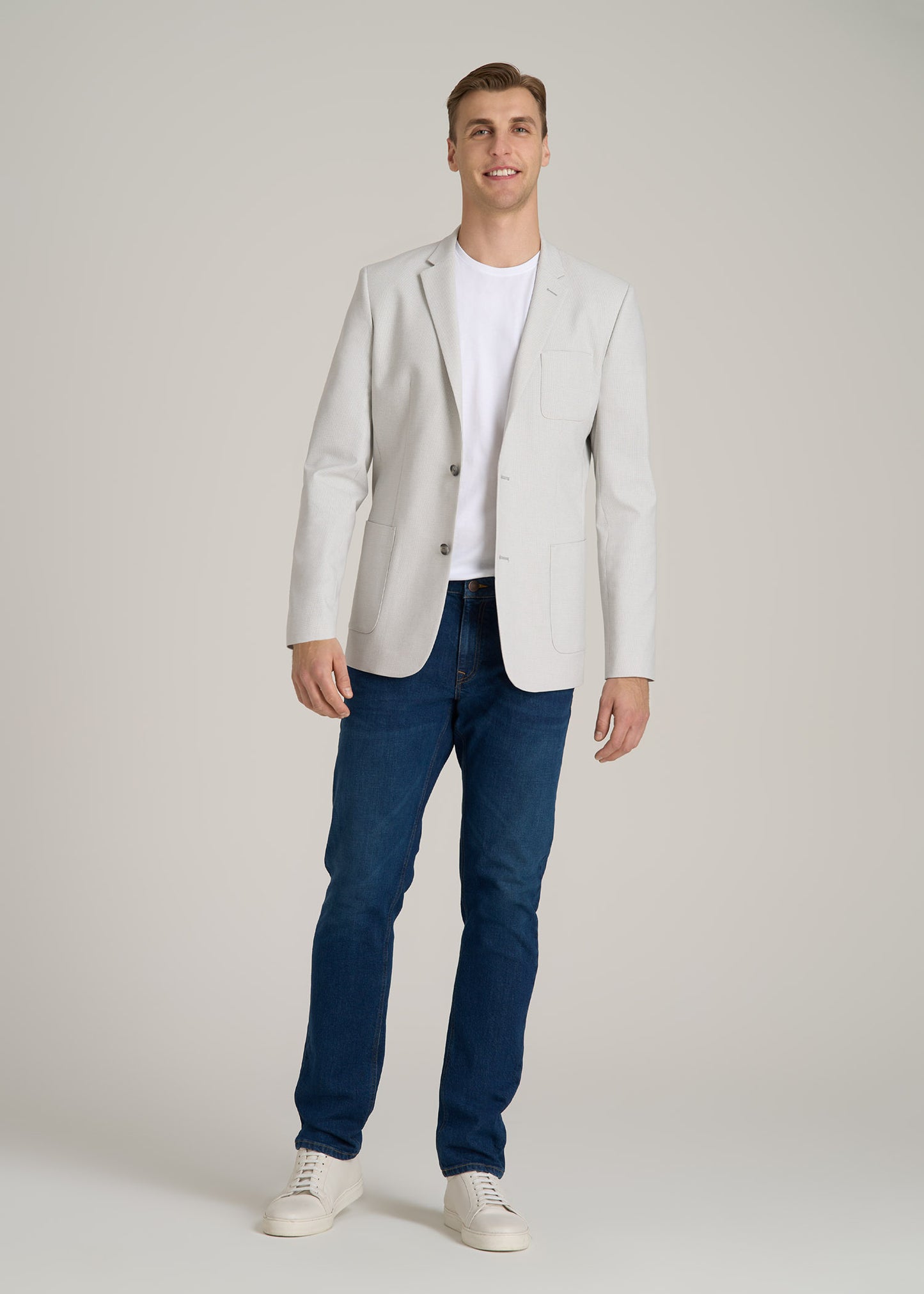 A tall man wearing American Tall's Men's Tall Blazer in Light Grey Houndstooth, Stretch Cotton MODERN-FIT T-Shirt for Tall Men in White, the Americana Collection Carman Tapered Fit Jeans For Tall Men in Crown Blue and Cupsole Tennis Sneakers for Tall Men in White.