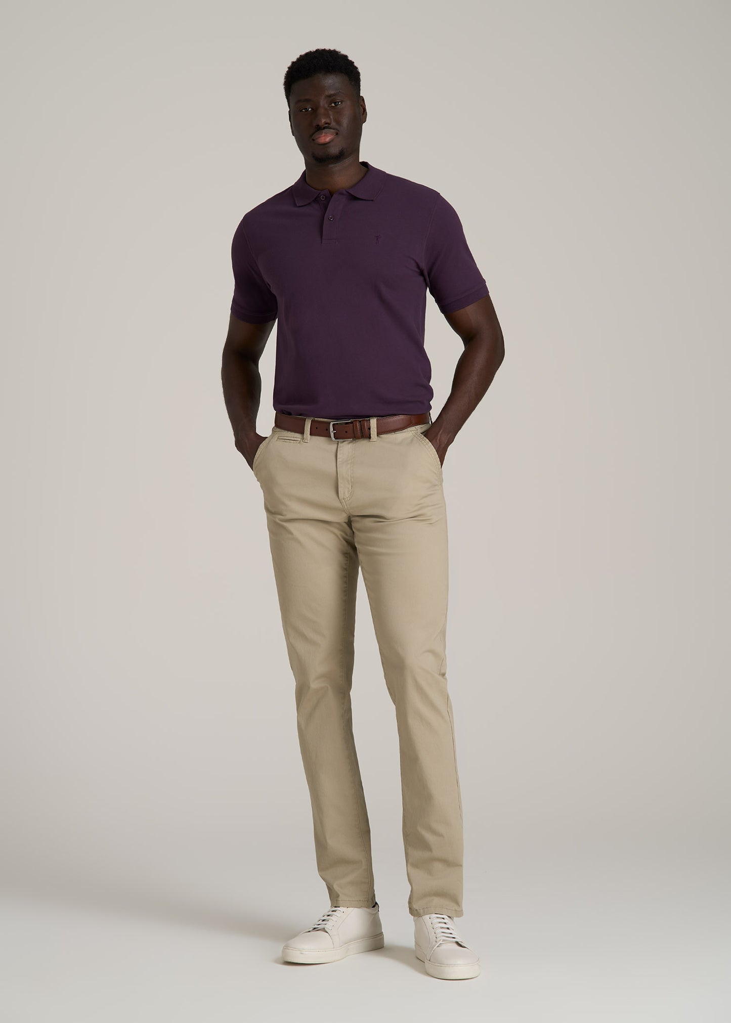 A tall man wearing American Tall's Men's Tall Classic Polo with Embroidered Logo in Midnight Plum, Carman TAPERED Chinos in Desert Khaki and Cupsole Tennis Sneakers for Tall Men in White.