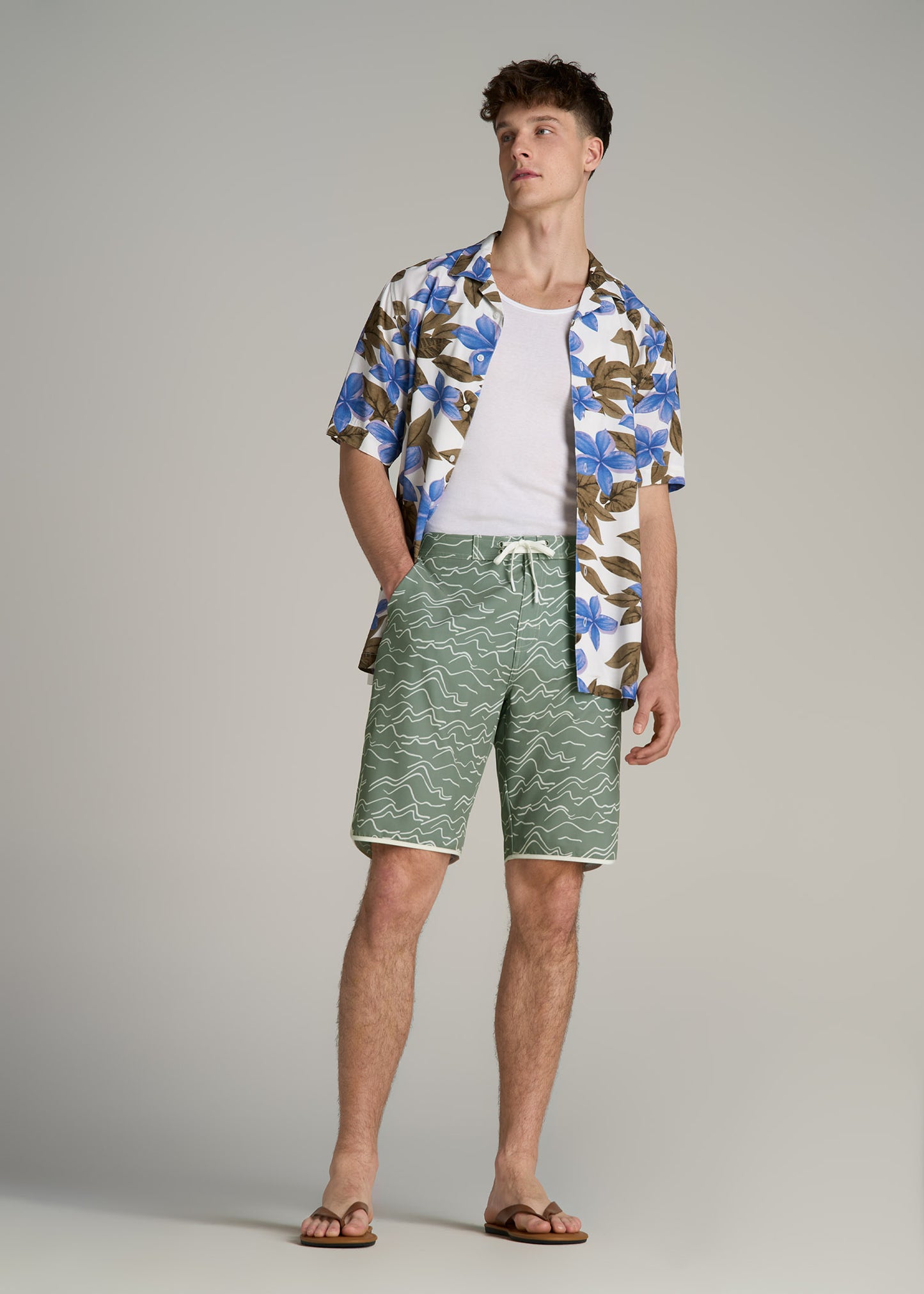 A tall man wearing American Tall's Hi-Tide Scallop Board Shorts in Green Current, Short Sleeve Resort Shirt in Blue Tropical Print and Men's Tall Ribbed Undershirt Tank Top in Bright White.