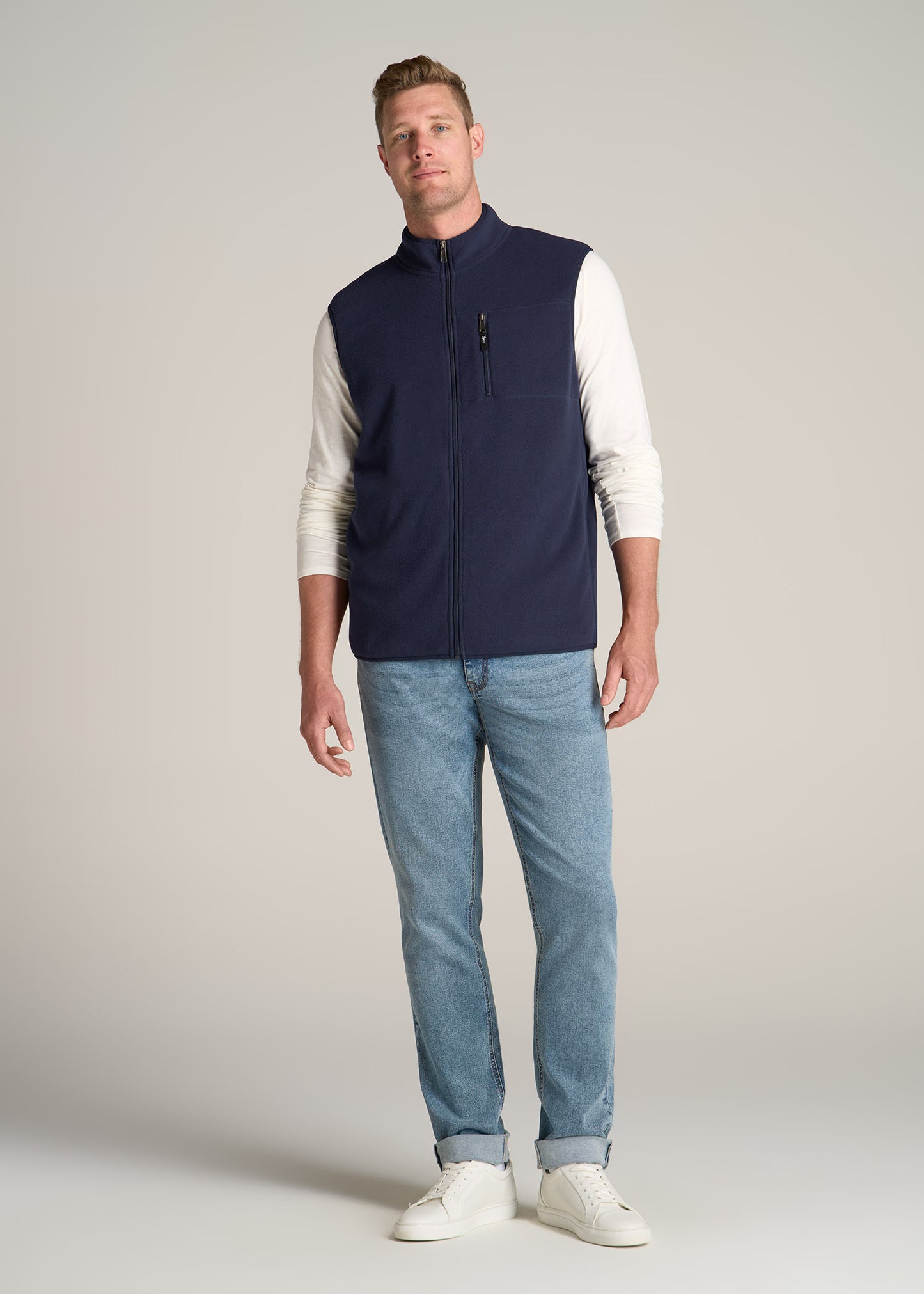 A tall man wearing American Tall's Polar Fleece Sweater Full Zip Vest for Tall Men in Regal Blue, their Slub Long Sleeve Scoop Tall Men's Tee in Ecru and Carman Tapered Jeans for Tall Men in Vintage Faded Blue.