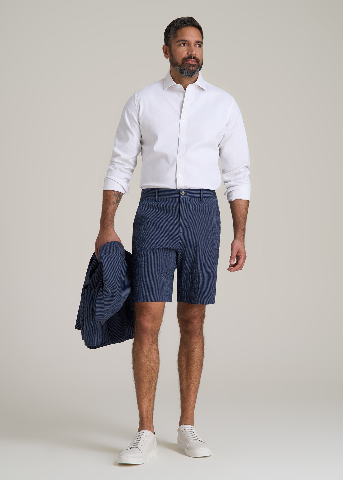 A tall man wearing American Tall's Seersucker Shorts in Navy and Off White Stripe and a
Stretch Linen Dress Shirt in White while holding a Stretch Seersucker Blazer in Navy and Off White Stripe.