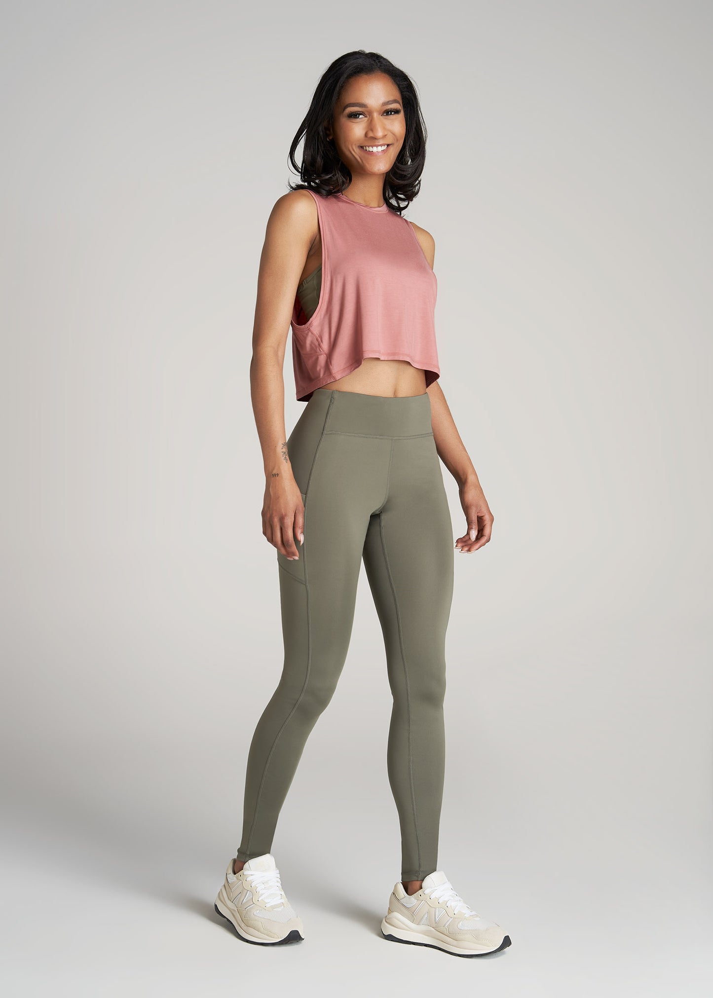 A tall woman wearing American Tall's Cropped Muscle Tank top in the color Clay Sunrise with olive green leggings.